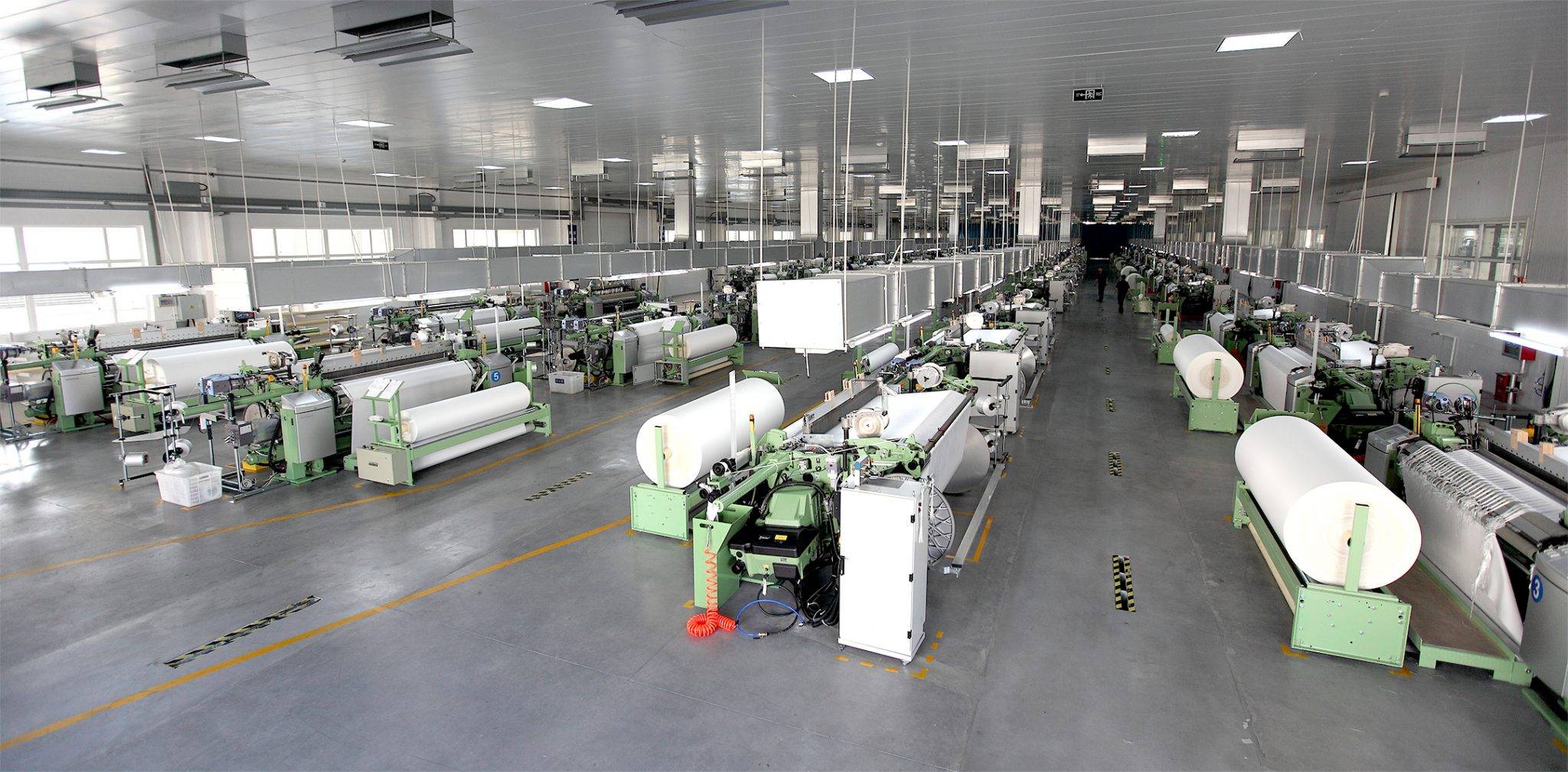 Spacious textile factory with rows of green weaving machines processing white fabric rolls