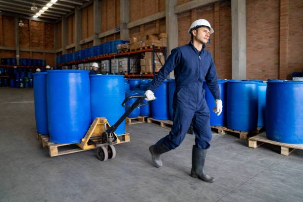 Worker in blue uniform moving blue chemical drums with a pallet jack in warehouse