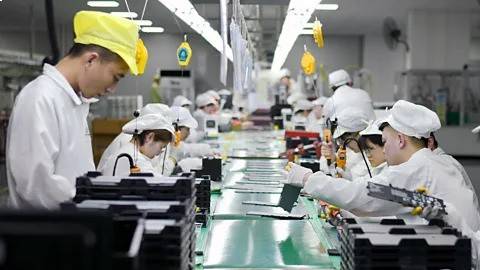 Asian Chinese factory workers in white uniforms and caps working on an automated battery production line in a modern facility.
