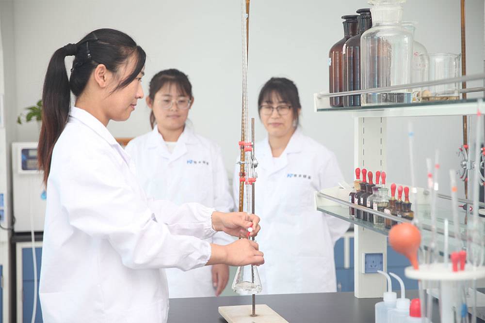 An Asian female chemist in a lab coat and safety glasses carefully inspecting a chemical sample in a test tube.